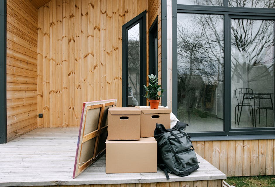 Inside a wooden-fronted home, a small collection of packing and moving items is arranged on a light-colored wooden porch next to large glass sliding doors. There are three cardboard boxes, two of which are stacked on top of each other, with a small potted plant placed on the top box. A black backpack and a cut piece of framed artwork with a wooden frame and fabric backing are leaning against the boxes. The sliding doors reveal a reflection of a garden with leafless trees and outdoor furniture, suggesting a move-in or home relocation process. The scene depicts the packing phase of a house move, with belongings prepared for transportation by Man with Van Wood Green, specialists in removals and furniture transport.