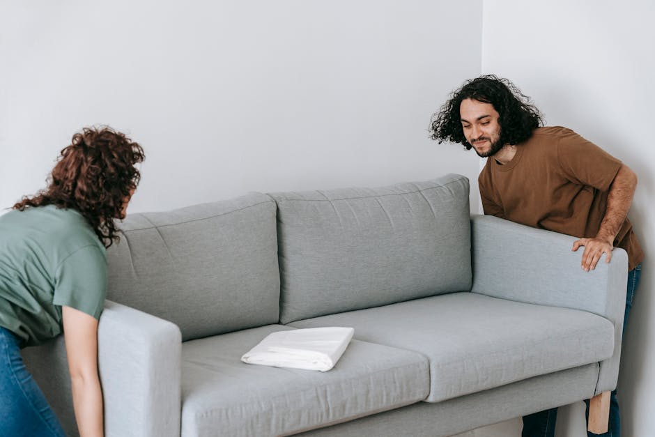 Two individuals are engaged in packing a light grey fabric-upholstered sofa with wooden legs inside a room, preparing it for a house move or furniture transport. The woman on the left, with curly red hair and wearing a green top, is securing the sofa with white packing blankets or protective covers to prevent damage during loading. The man on the right, with long dark curly hair, a beard, and wearing a brown T-shirt, is holding the armrest of the sofa, assisting in the packing or lifting process. The setting appears to be a well-lit interior space with white walls and minimal decor, indicating a typical home environment. On the sofa's seat, there are folded white protective fabrics or blankets, commonly used in packing and moving to safeguard furniture. This scene relates to the process of home relocation, specifically lifting and packing furniture for transportation, with no vehicles or additional equipment visible. Man with Van Wood Green specializes in removals, including furniture handling and loading for house moves and large item transportation.