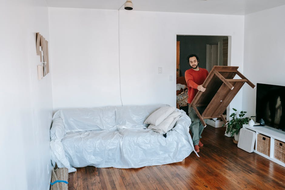 A man moving a wooden dining table inside a living room during a home removal process. The room features white walls, a hardwood floor, and minimal furniture, including a white sofa covered with a protective plastic sheet and a few cushions. The man, dressed in casual clothing with a red top and green pants, is holding the table by its sides, preparing to load it onto a vehicle or move it across the room. Nearby, there is a flat-screen television placed on a white shelving unit with woven baskets, and a potted plant on the floor. A doorway in the background reveals another room, and natural light illuminates the scene. This image, associated with Man with Van Wood Green, illustrates furniture transport and packing during a typical house relocation or flat move, emphasizing careful handling and organization during the loading process for an efficient removals service.