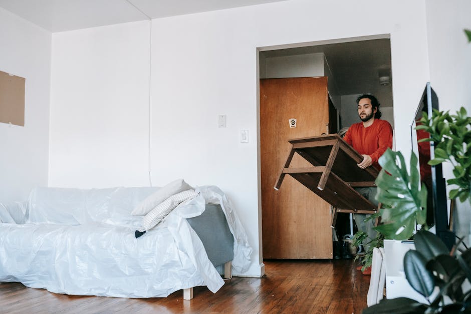 Two individuals are engaged in packing a light grey fabric-upholstered sofa with wooden legs inside a room, preparing it for a house move or furniture transport. The woman on the left, with curly red hair and wearing a green top, is securing the sofa with white packing blankets or protective covers to prevent damage during loading. The man on the right, with long dark curly hair, a beard, and wearing a brown T-shirt, is holding the armrest of the sofa, assisting in the packing or lifting process. The setting appears to be a well-lit interior space with white walls and minimal decor, indicating a typical home environment. On the sofa's seat, there are folded white protective fabrics or blankets, commonly used in packing and moving to safeguard furniture. This scene relates to the process of home relocation, specifically lifting and packing furniture for transportation, with no vehicles or additional equipment visible. Man with Van Wood Green specializes in removals, including furniture handling and loading for house moves and large item transportation.
