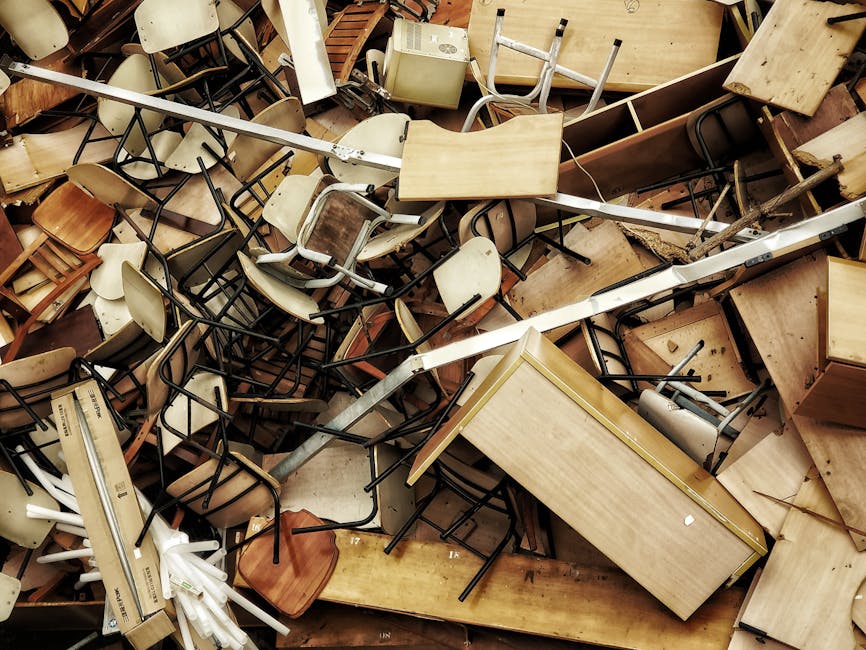A collection of outdoor furniture and packaging materials arranged on a paved surface next to a stone wall, including wooden chairs in various shades of natural wood, painted or treated in grey and bl