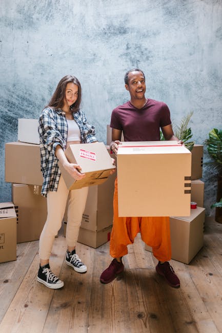 A young woman and a man are standing indoors on a wooden floor against a textured concrete wall, each holding medium-sized cardboard boxes filled with items. The woman, dressed casually in a checkered shirt, white top, beige pants, and black-and-white sneakers, is smiling and facing slightly to the side. The man, wearing a maroon T-shirt, orange trousers, and burgundy shoes, is also smiling and looking directly at the camera. Several additional cardboard boxes are stacked behind them, some sealed and others open, suggesting they are in the process of a home relocation or moving activity. The scene includes indoor plant decor on the right side, with natural lighting illuminating the space. This image illustrates packing, handling, and the logistics involved in furniture transport and house moves, relevant to the context of house removals and compliance with regulations like Haringey Council’s bulky-item rules, as covered by Man with Van Wood Green.