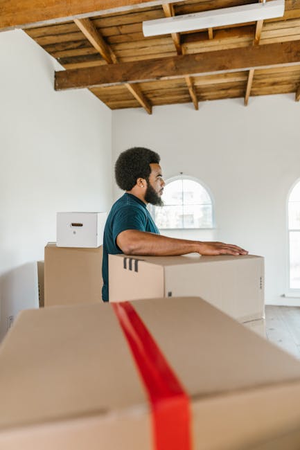 A man with dark curly hair and a beard, dressed in a teal shirt, is standing inside a bright, empty room with white walls and a wooden ceiling with exposed beams. He is positioned behind a large cardboard box, with his arms resting on top of it, participating in a home relocation or furniture transport process. Several other cardboard boxes of varying sizes, some sealed with red or white tape, are stacked around him, indicating packing and moving preparations. The room has two arched windows allowing natural light to illuminate the scene. Visible outside the windows are blurred buildings. The image captures the loading or unpacking stage of house removals, with furniture and boxes inside the property, possibly being prepared for transport. This scene is associated with services such as those offered by Man with Van Wood Green, focusing on efficient, professional moving solutions, including packing, lifting, and vehicle loading, relevant to local council regulations on bulky-item disposal and house removals.