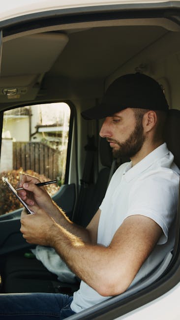 A small maroon flatbed van parked on a city street outside a modern building with large glass windows and a concrete facade. The van's open cargo area is filled with various packed boxes wrapped in pl