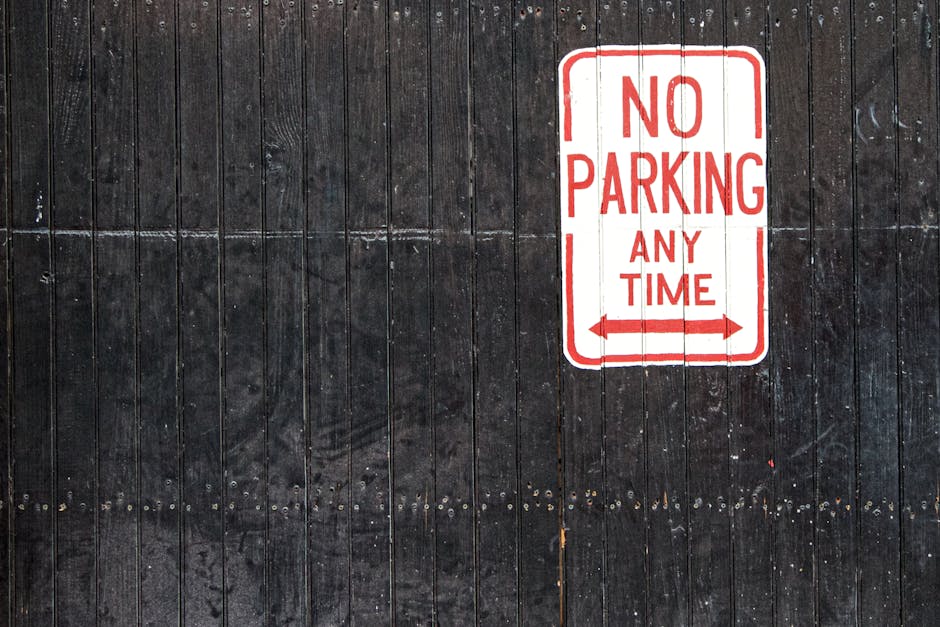 A close-up image of a rectangular 'No Parking Any Time' sign with red text and arrows pointing left and right, mounted on a dark, weathered wooden fence with vertical planks. The surface of the fence shows visible grain and texture, with some small nails or rivets along the bottom edge. The sign is situated in an outdoor environment, likely at the entrance or boundary of a property, indicating parking restrictions. This setting relates to house removals and moving logistics, where adherence to parking restrictions is essential for a smooth loading process, as managed by Man with Van Wood Green. The lighting is natural, highlighting the contrast between the white sign and dark background, emphasizing the importance of parking regulations during home relocation or furniture transport activities.