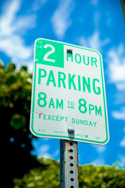 A vertical green and white parking restriction sign mounted on a metal pole outdoors, indicating a two-hour parking limit from 8 am to 8 pm, except on Sundays. The sign features bold green lettering and numbers, with smaller details including the cost and jurisdiction at the bottom. The background includes a clear blue sky with some scattered clouds and blurred green foliage, suggesting a residential or urban street setting. This image is relevant to home relocation and furniture transport services offered by Man with Van Wood Green, illustrating parking regulations that may impact the logistics of loading and unloading during house removals or moving services.