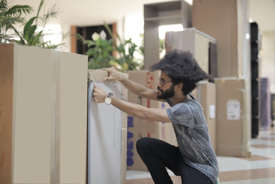 A man with curly black hair, glasses, and a beard is kneeling inside a spacious, well-lit indoor environment, preparing to load large cardboard boxes wrapped in packing paper onto a trolley for a move. He is wearing a patterned grey shirt, black trousers, and a wristwatch. The boxes vary in size and are positioned on the floor close to a doorway or open area, with some stacked in the background. The scene shows a professional furniture transport and packing process, with the man carefully securing or arranging the boxes as part of a home or office relocation handled by Man with Van Wood Green. In the background, there are shelves, potted plants, and a television, indicating a domestic or commercial setting awaiting transition. The overall environment appears organized, with the focus on safe handling of household or office items for moving services, utilizing packing materials and equipment such as trolleys to facilitate the loading process.