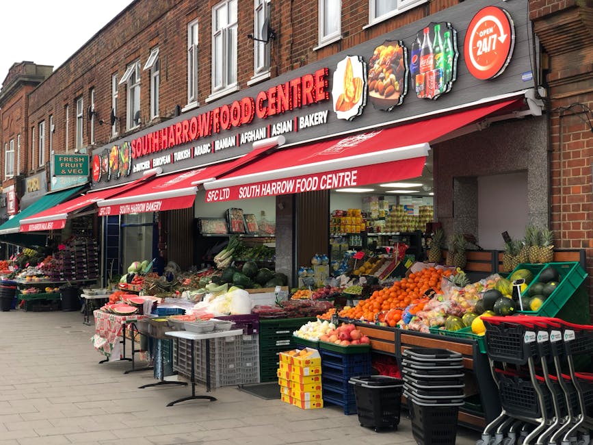 The image shows the exterior of South Harrow Food Centre, a local grocery with a red and white sign displaying the store name and various food categories such as bakery, Asian, Afghan, and Turkish products. The storefront features a red and white striped awning extending over the sidewalk, with additional signage indicating 24/7 opening hours. Outside, there are multiple vendor tables and green crates arranged along the pavement, loaded with fresh produce including oranges, apples, watermelons, cucumbers, and other fruits and vegetables. Some items are wrapped in plastic or placed in baskets, with an assortment of packaging materials visible. Several black plastic crates and shopping carts are stacked nearby, ready for transport or delivery. The scene captures activity typical of a busy food shop during daylight, with natural lighting illuminating the produce and storefront, suggesting a context of packing, loading, or preparing for home or business relocations. Occasionally, Man with Van Wood Green’s services might be involved in removals or transport, supporting clients with moving and logistics through professional furniture transport and packing processes.