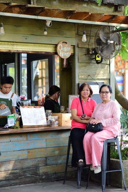 The image shows a small outdoor seating area of a cafe or food shop, with a rustic wooden exterior wall painted in pastel shades of blue and green. There is a window opening with a counter where a sta