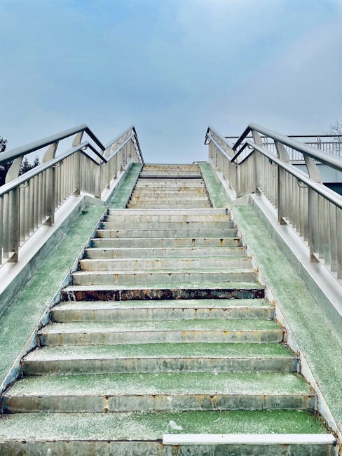 A set of outdoor concrete stairs with metal railings on both sides, leading up to an elevated platform or rooftop. The stairs are weathered, with visible signs of wear and patches of moss or algae on the surface. The railing sections are metallic and painted in a dark color, with horizontal bars providing support. The sky above is overcast, with a cloudy grey tone, indicating a dull weather day. This scene could be part of a residential or commercial building, illustrating the type of access involved in home relocations or furniture transport. [COMPANY_NAME] may assist with navigating such access points during house removals or moving services, ensuring safe and efficient furniture transport and packing during a home relocation.