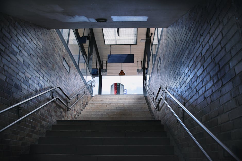 Interior view of a staircase within a brick-walled building, leading upwards to an outdoor area with daylight visible at the top. The staircase has dark, wide steps with metal handrails on both sides. At the top of the stairs, there are glass panels and a small outdoor corridor with a red and grey structure in the background. The scene appears to be part of a house removal or moving process, with the location being prepared for furniture transport. The lighting in the stairwell is primarily natural, highlighting the textured brick walls and metal railings, supporting the context of efficient access and movement logistics, as referenced in the page about Turnpike Lane removals by Man with Van Wood Green.
