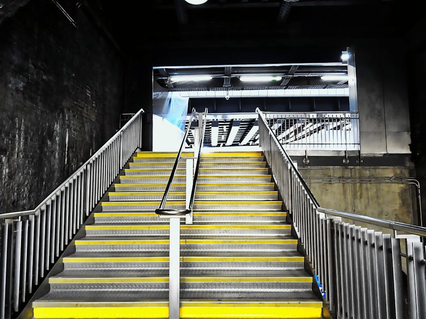 A set of outdoor concrete stairs with metal railings on both sides, leading up to an elevated platform or rooftop. The stairs are weathered, with visible signs of wear and patches of moss or algae on the surface. The railing sections are metallic and painted in a dark color, with horizontal bars providing support. The sky above is overcast, with a cloudy grey tone, indicating a dull weather day. This scene could be part of a residential or commercial building, illustrating the type of access involved in home relocations or furniture transport. [COMPANY_NAME] may assist with navigating such access points during house removals or moving services, ensuring safe and efficient furniture transport and packing during a home relocation.
