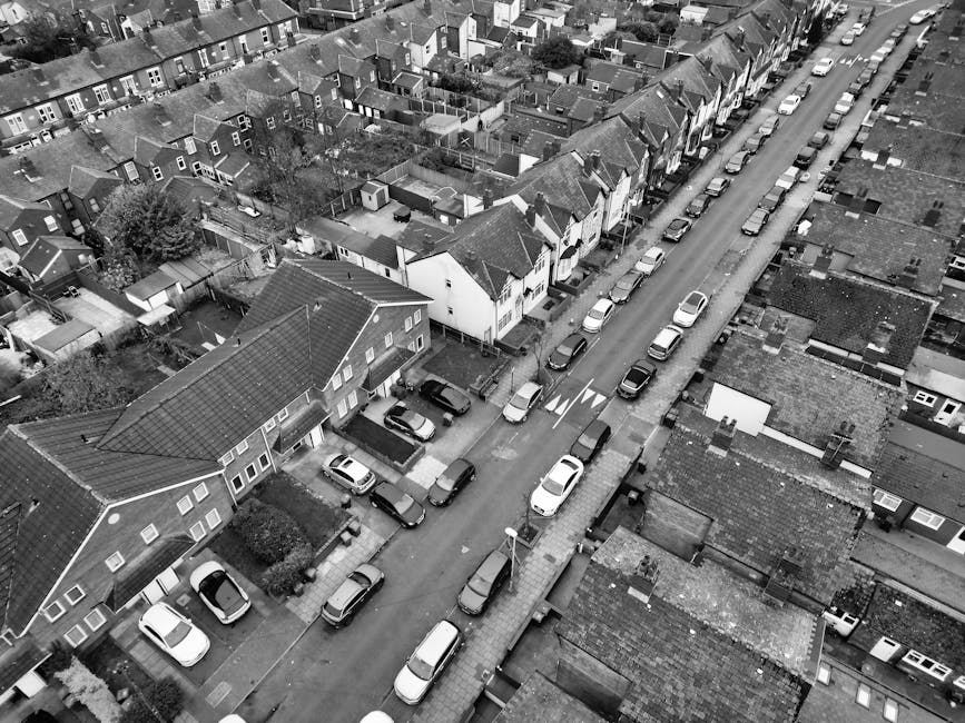 A black and white aerial photograph of a residential street in Wood Green, showing a line of parked cars along both sides of the road, which is flanked by terraced houses with pitched roofs and small front gardens. In the foreground, a house with a tiled roof is visible, with several cardboard boxes and wrapped furniture pieces on the pavement near the entrance, likely part of a home relocation process. A man with a van from Man with Van Wood Green is partially visible, loading or unloading large furniture items covered in blankets and secured with straps into a white van parked on the street. The scene captures the logistical planning involved in furniture transport and packing during a house removal, with an emphasis on careful handling of household items in a neighborhood setting during a moving and loading process. The environment is well-lit, with natural daylight illuminating the scene and highlighting the organized approach to residential removals on narrow streets.